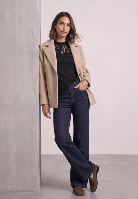 Eine Frau lehnt an einer Säule, trägt beige Blazer, schwarze Spitzenbluse und dunkle Jeans.