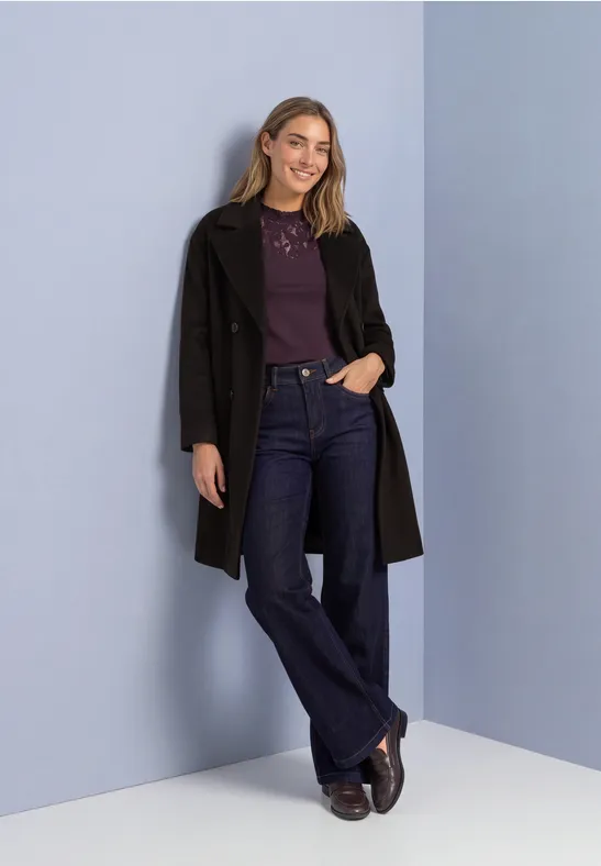 Frau lehnt an pastellblauer Wand, trägt schwarzen Mantel, violettes Oberteil, weite Jeans und braune Schuhe.