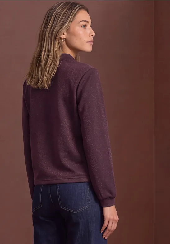 Seitlich stehende Frau in bordeauxrotem Pullover und Jeans vor braunem Hintergrund.