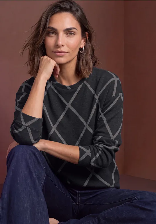Femme portant un pull sombre à motifs géométriques, assise de manière décontractée devant un fond neutre.