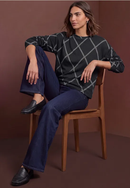 Femme assise sur une chaise, porte un pull gris foncé à motifs quadrillés blancs, jean bleu foncé et mocassins noirs.