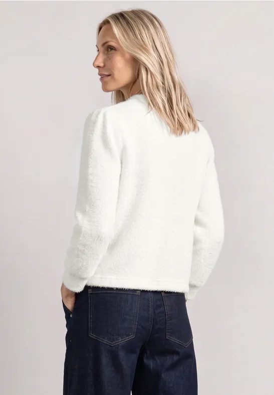 Eine blonde Frau von hinten in weißer Jacke und blauen Jeans vor hellem Hintergrund.