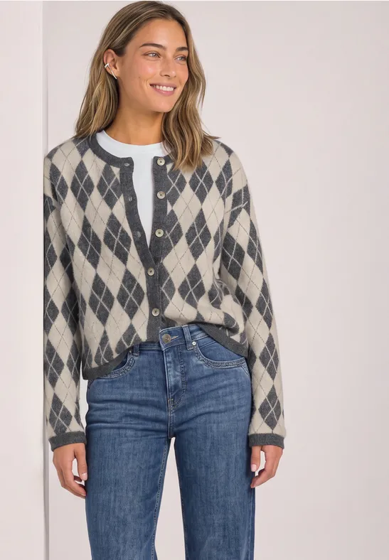 Eine Frau lächelt und trägt einen graubeigen Cardigan mit Rautenmuster, darunter ein weißes Shirt und blaue Jeans.