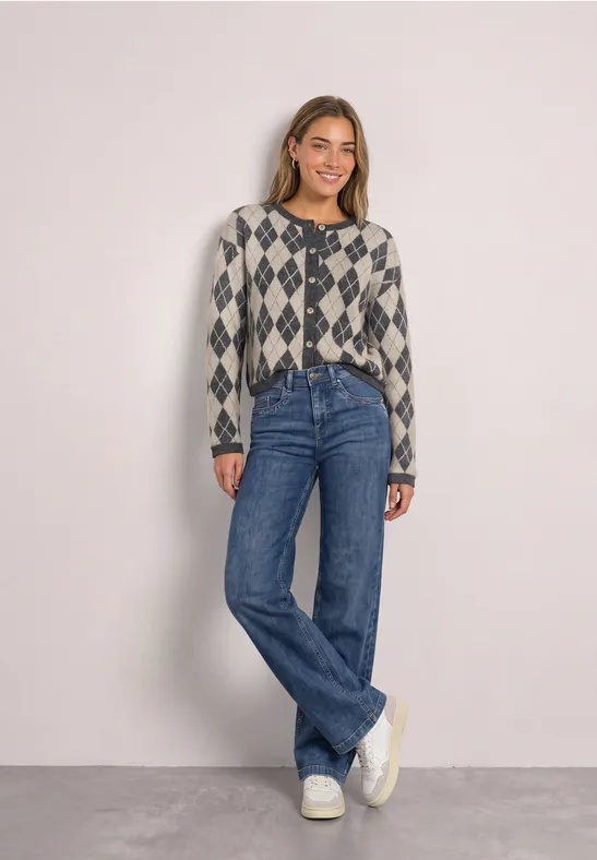Frau in karierter Strickjacke, blauen Jeans und weißen Sneakers vor schlichtem Hintergrund.