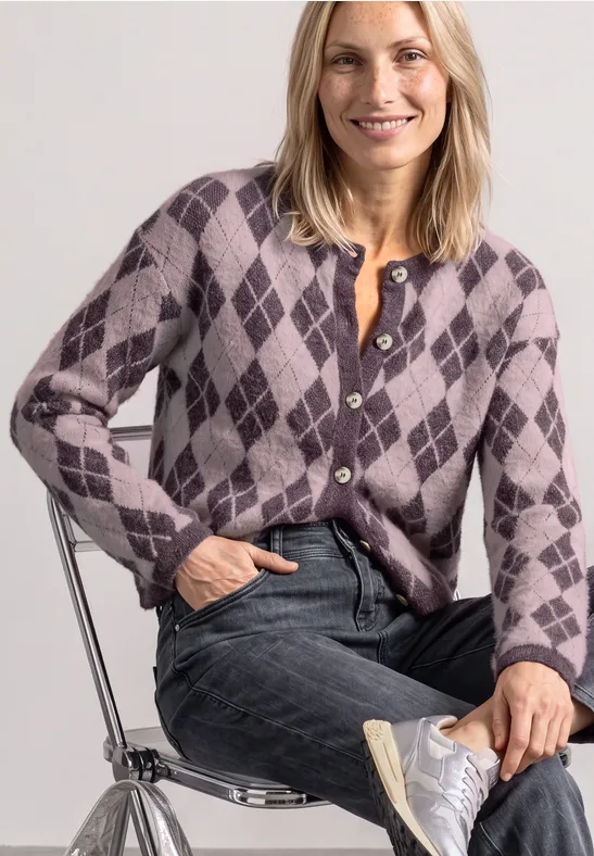 Frau im violetten Rautenmuster-Cardigan, Jeans und weißen Sneakern, lächelnd auf einem Stuhl sitzend.