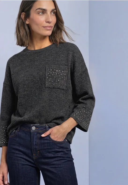 Frau in dunkelgrauem Pullover mit funkelnder Tasche und dunklen Jeans vor hellem Hintergrund.