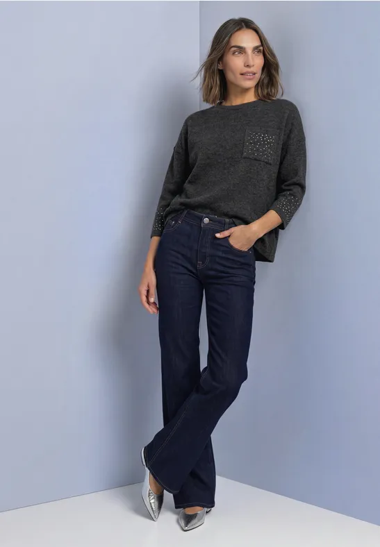 Frau lehnt an Wand, trägt grauen Pullover, dunkelblaue Schlagjeans und silberne Schuhe.