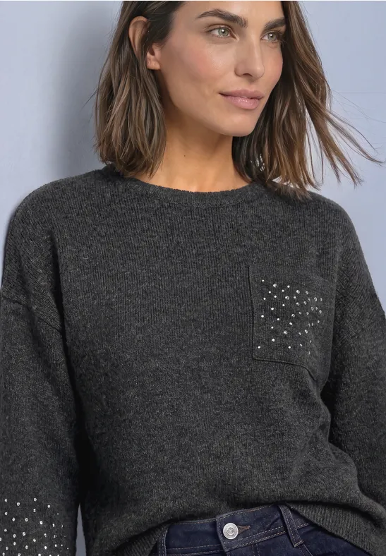 Frau in grauem Strickpullover mit Nieten auf der Tasche und Jeans, schaut seitlich.