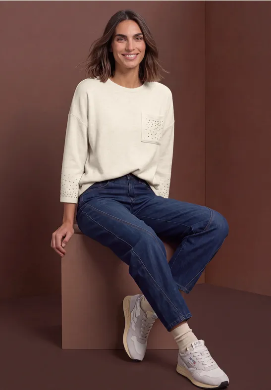 Eine Frau in lässiger Kleidung: helle 3/4-Arm-Bluse, dunkle Jeans und weiße Sneakers vor braunem Hintergrund.