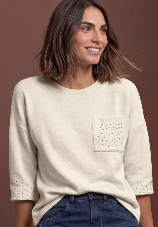 Eine lächelnde Frau mit braunen Haaren trägt einen hellbeigen Pullover mit dekorativer Brusttasche.