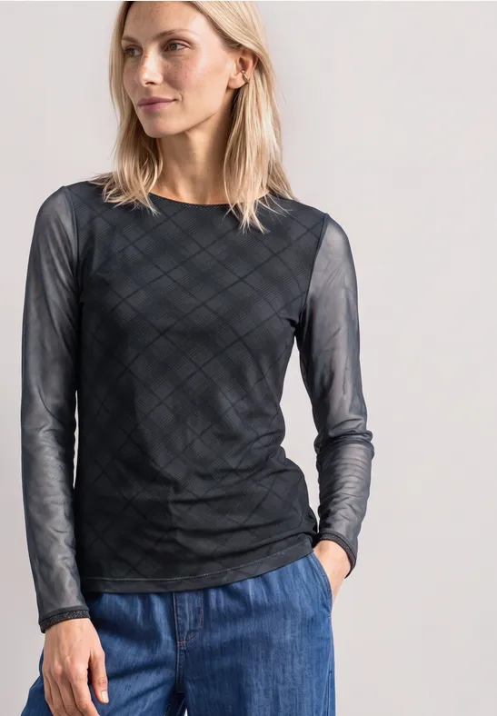 Frau in durchsichtigem schwarzen Langarmshirt mit Karomuster und blauer Jeans vor hellem Hintergrund.