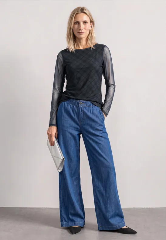 Eine Frau in schwarzem Mesh-Oberteil, weiten Jeans und schwarzen Schuhen mit silberner Clutch.