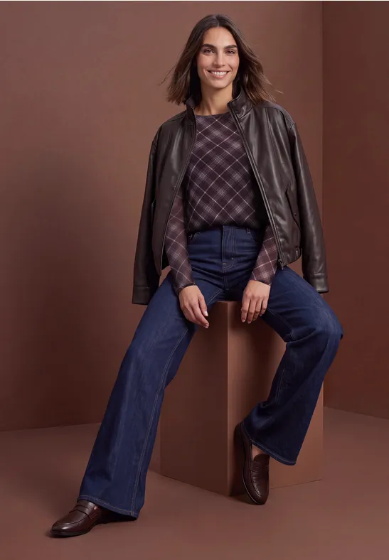 Vrouw met leren jas over schouders, ruitjestrui, hoge flare-jeans en bruine enkellaarsjes.