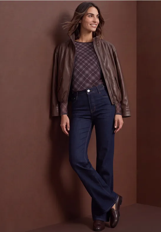 Vrouw met bruine leren jas, gebreide trui, donker flared jeans en laarzen voor een bruine achtergrond.