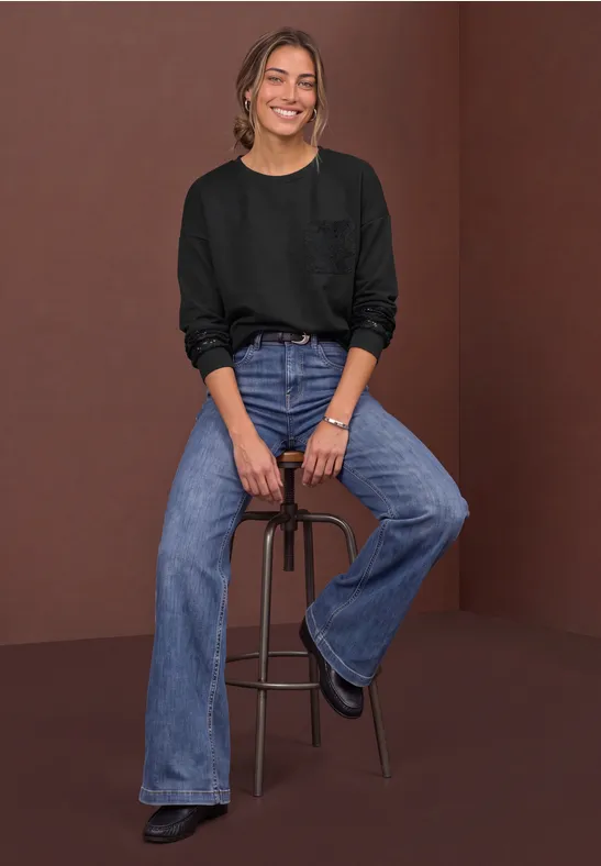 Junge Frau in schwarzem Pullover und weiten Jeans sitzt lächelnd auf einem Hocker vor braunem Hintergrund.