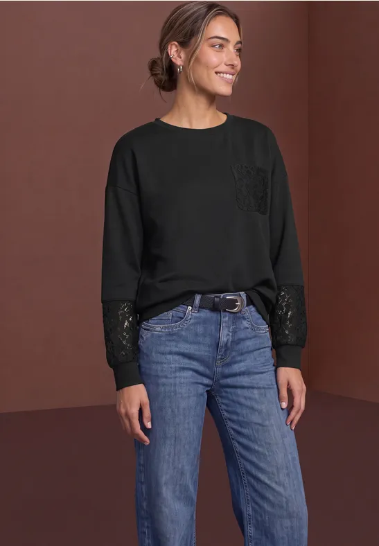 Frau in schwarzem Sweatshirt mit Paillettenärmeln und Jeans steht vor braunem Hintergrund und lächelt.