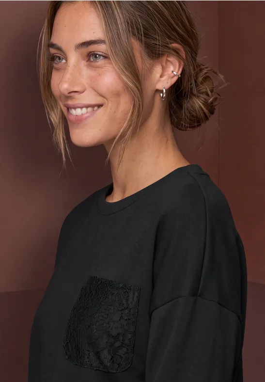 Junge Frau mit zurückgebundenem Haar, lächelnd, trägt schwarzen Pullover mit Spitzentasche.