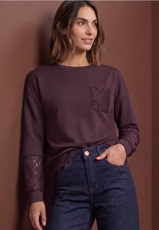 Frau in lila Langarmshirt mit Spitzenbesatz an Tasche und Ärmeln, dunkle Jeans, vor braunem Hintergrund.