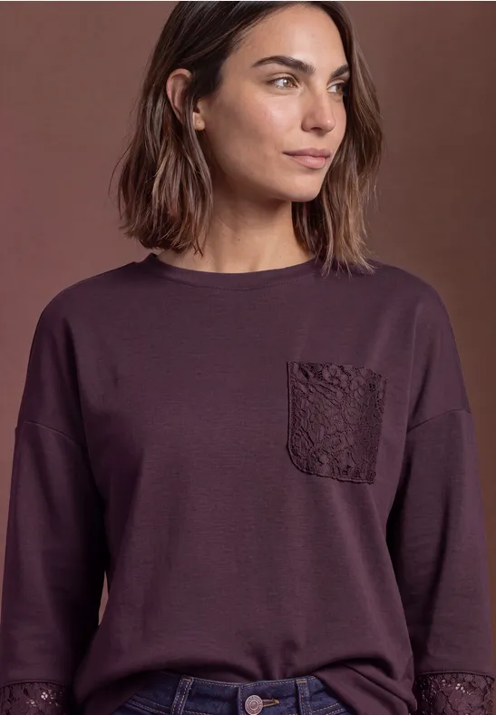 Frau mit schulterlangem Haar trägt ein dunkelviolettes Langarmshirt mit dekorativer Brusttasche.