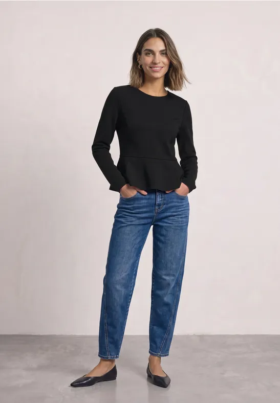 Frau in schwarzem Langarmshirt, blauen Jeans und Ballerinas lächelt mit Händen in den Taschen vor hellem Hintergrund.