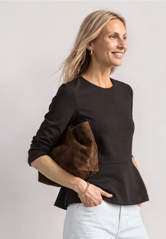 Frau in schwarzem Peplum-Top und weißen Hosen, lächelt und hält eine braune Clutch vor hellem Hintergrund.
