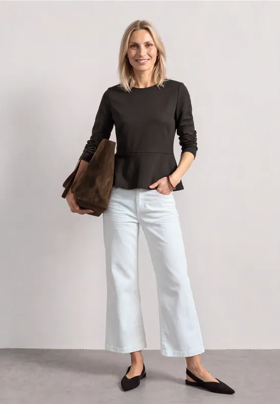 Eine Frau vor hellem Hintergrund trägt dunkelgraues Oberteil, weiße weite Hosen, schwarze Ballerinas, braune Tasche.