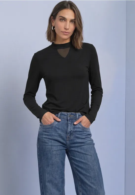 Frau in schwarzem Langarmshirt mit transparentem Einsatz am Ausschnitt und blauer Jeans, Hände in Taschen.