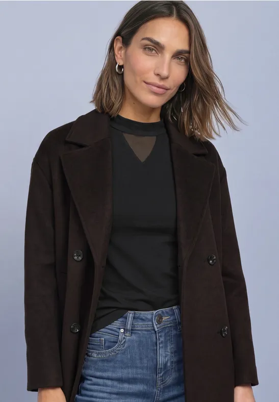 Junge Frau in schwarzem Top, dunklem Mantel und Jeans vor neutralem Hintergrund.