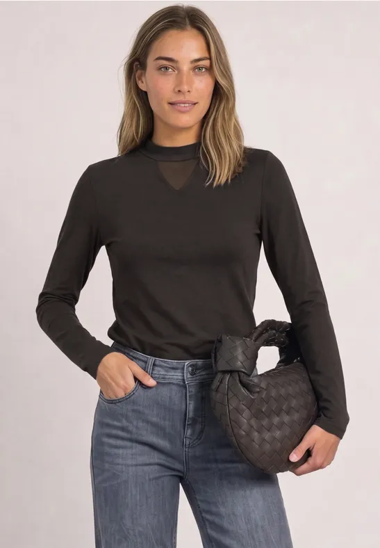 Junge Frau trägt schwarzes Langarmshirt, Jeans und eine gesteppte schwarze Handtasche vor hellem Hintergrund.