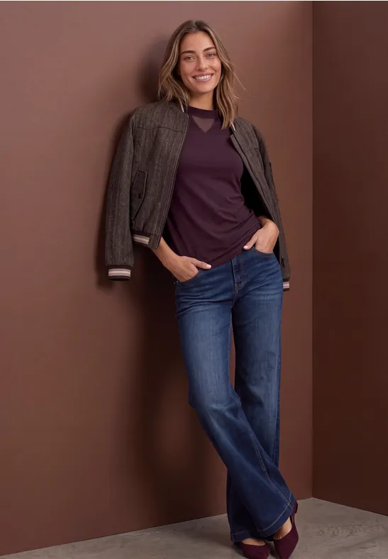 Frau lehnt an brauner Wand, trägt Jeans, violettes Shirt und Jacke über den Schultern, lächelt.