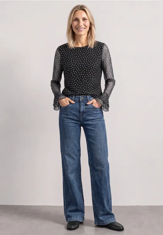 Frau in schwarz-weiß gepunkteter Bluse mit Rüschenärmeln und weiten blauen Jeans schwarze Schuhe vor grauem Hintergrund.