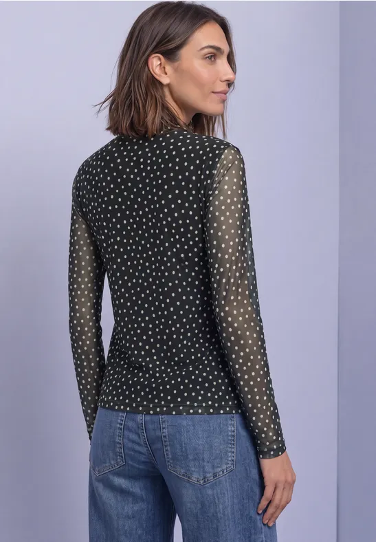 Frau im Profil mit schwarzem, gepunktetem Mesh-Langarmshirt und blauer Jeans.