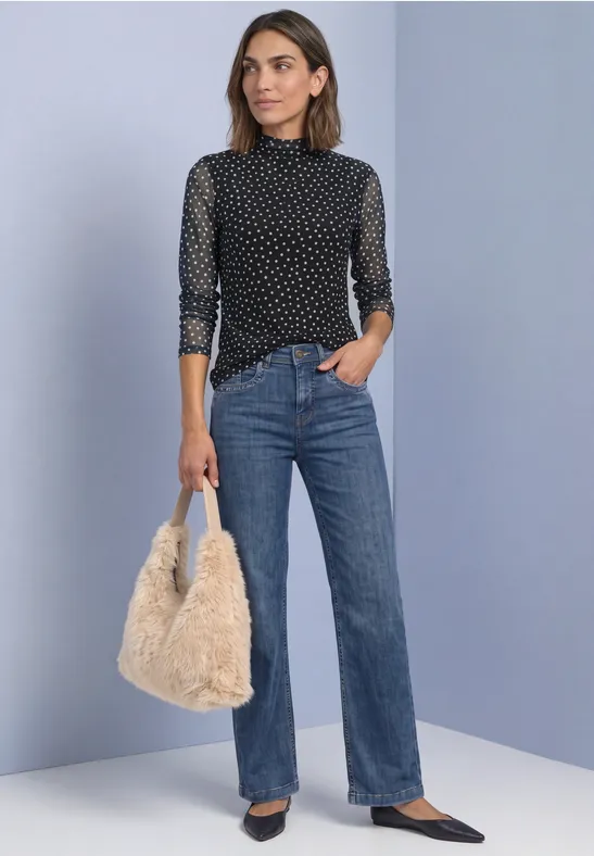 Frau mit schwarzer, gepunkteter Bluse, blauen Jeans und flauschiger Tasche vor schlichtem Hintergrund.