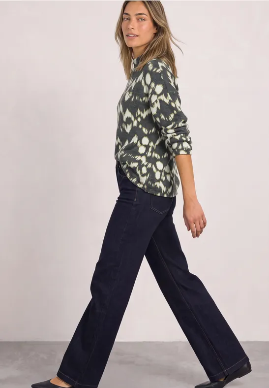 Frau in grüner, gemusterter Bluse und weiten Jeans in seitlicher Pose vor hellem Hintergrund.