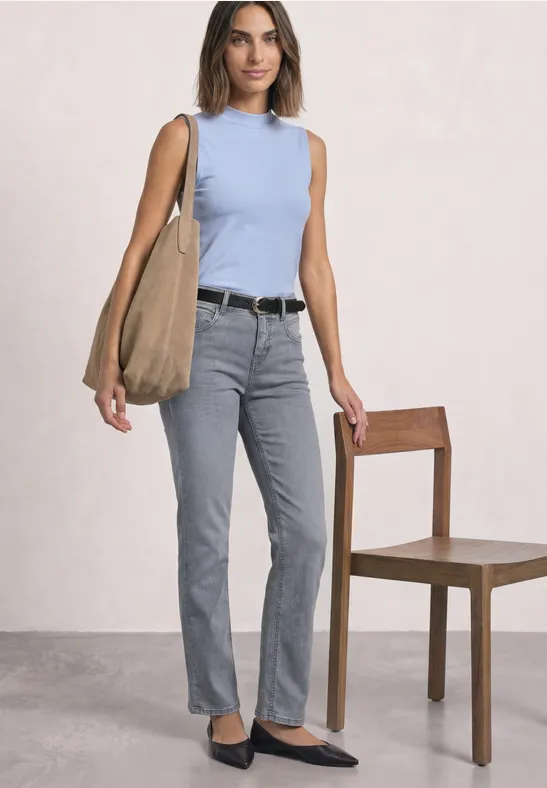 Frau trägt hellblaues Top, graue Jeans, schwarze Ballerinas und große Schultertasche vor hellem Hintergrund.