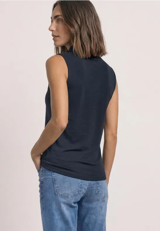 Frau in ärmellosem dunkelblauen Top und Jeans, seitlich stehend, Hand in Tasche.