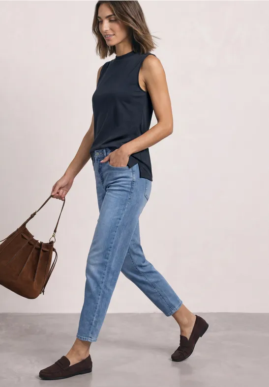 Frau in schwarzem Top, blauen Jeans und schwarzen Slippern trägt braune Tasche und geht.