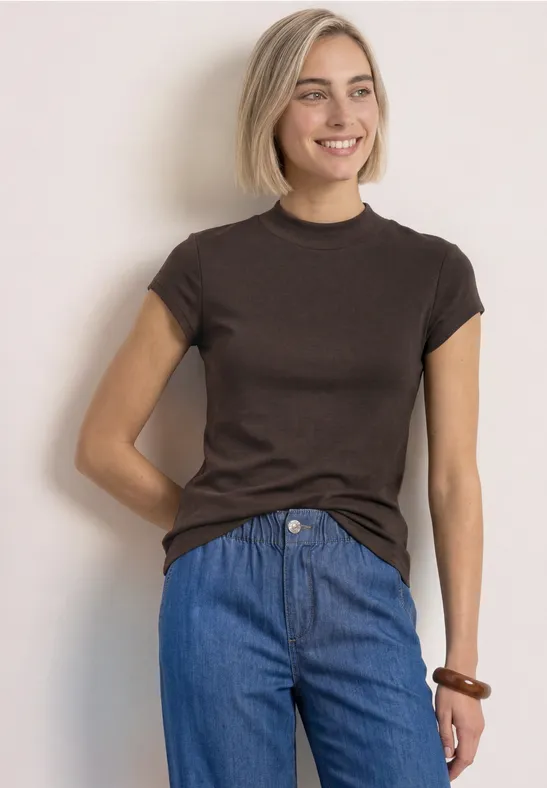 Eine lächelnde Frau in braunem T-Shirt und blauen Jeans vor hellem Hintergrund.