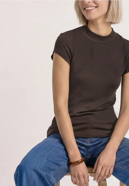 Frau sitzt lächelnd, trägt braunes T-Shirt, blaue Jeans und Holzarmband.