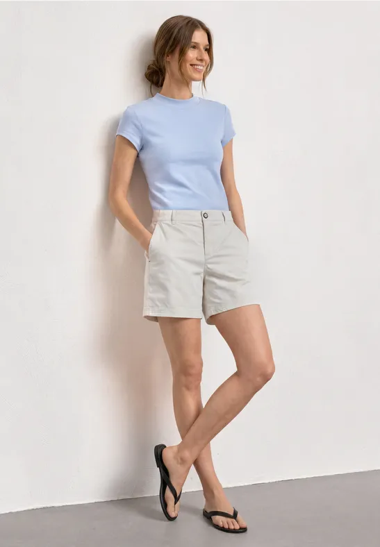 Frau in hellblauem T-Shirt, beigen Shorts und schwarzen Flip-Flops lehnt lässig an weißer Wand.