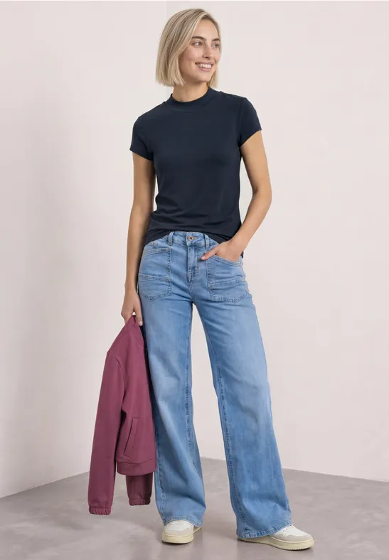 Frau trägt schwarzes T-Shirt, weite Jeans und weiße Sneakers, hält eine rosa Jacke.