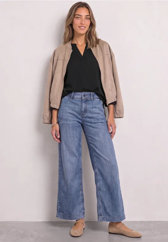 Frau in lockerer Jeans mit weitem Bein, schwarzem Top und beiger Jacke, lässig mit hellen Slippern.