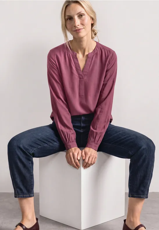 Frau sitzt auf weißem Würfel, trägt violette Bluse und Jeans, entspannt und lässig.