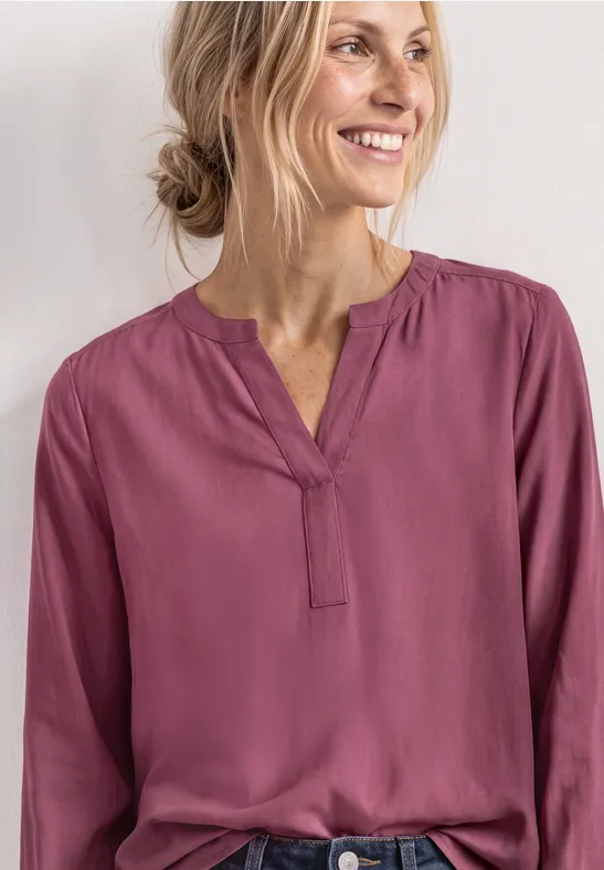 Eine lächelnde Frau in einer violetten, locker sitzenden Bluse vor hellem Hintergrund.