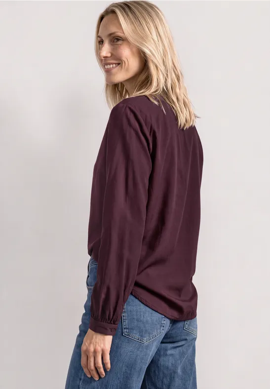 Eine Frau mit lockeren blauen Jeans und einer dunkellila Bluse schaut über die Schulter und lächelt.