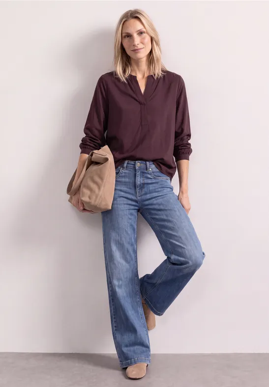 Eine Frau in violetter Bluse und weiten Jeans mit Beuteltasche vor neutralem Hintergrund.