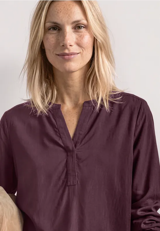 Lächelnde, blonde Frau in einem bordeauxroten Oberteil vor hellem Hintergrund.