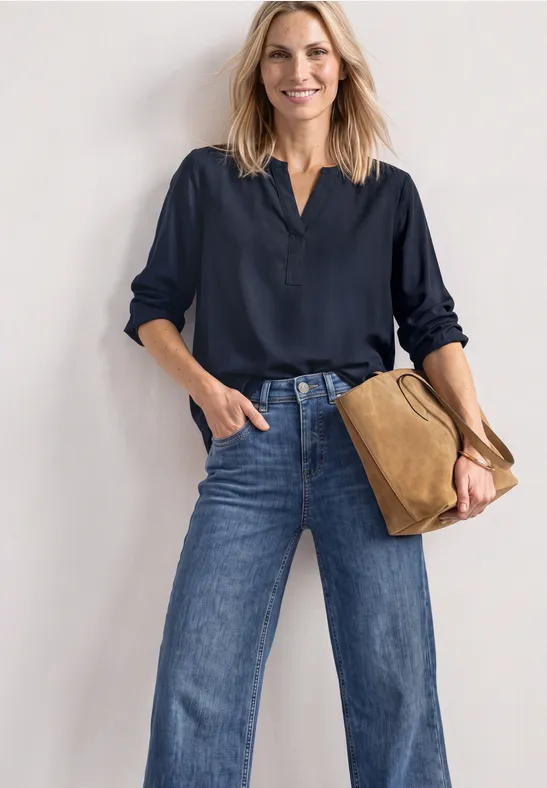 Frau in dunkelblauer Bluse und weitem Jeans, mit brauner Tasche, steht lächelnd vor heller Wand.