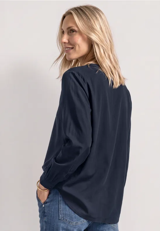 Eine Frau mit blonden Haaren trägt eine dunkelblaue Bluse und Jeans, schaut lächelnd über die Schulter.