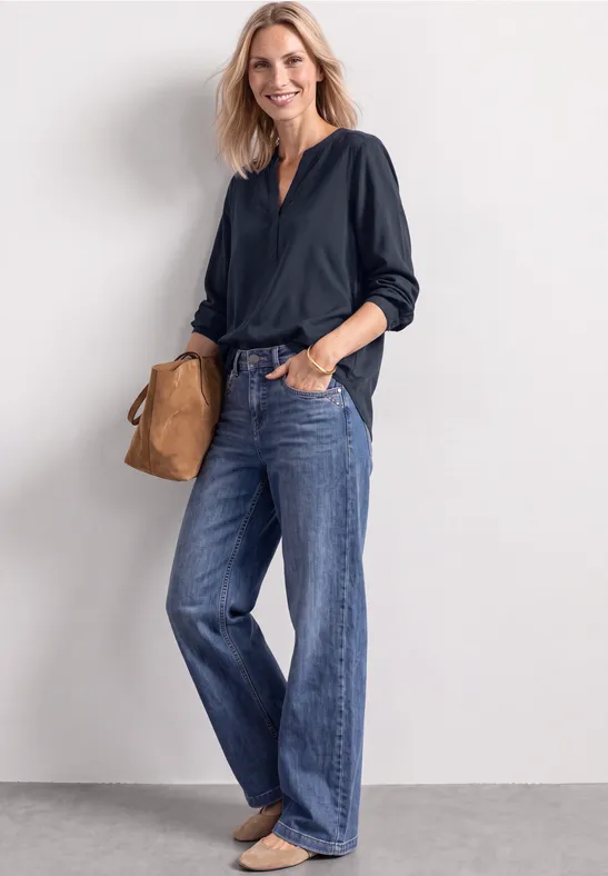 Frau in dunkler Bluse und weitem Jeans vor hellem Hintergrund mit brauner Tasche.